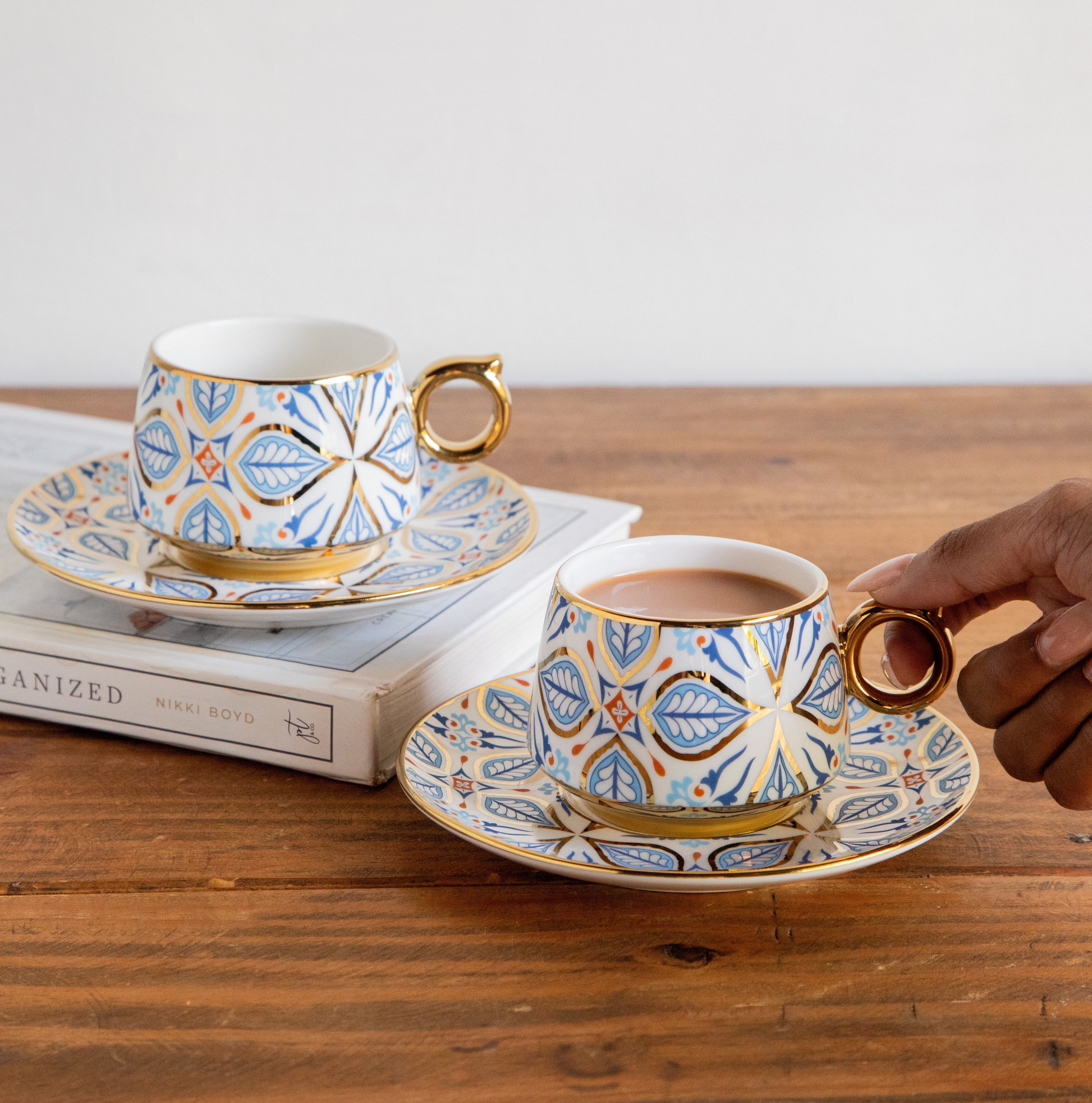 Decorative tea cup and saucer set on a wooden table with a hand holding one of the cups.