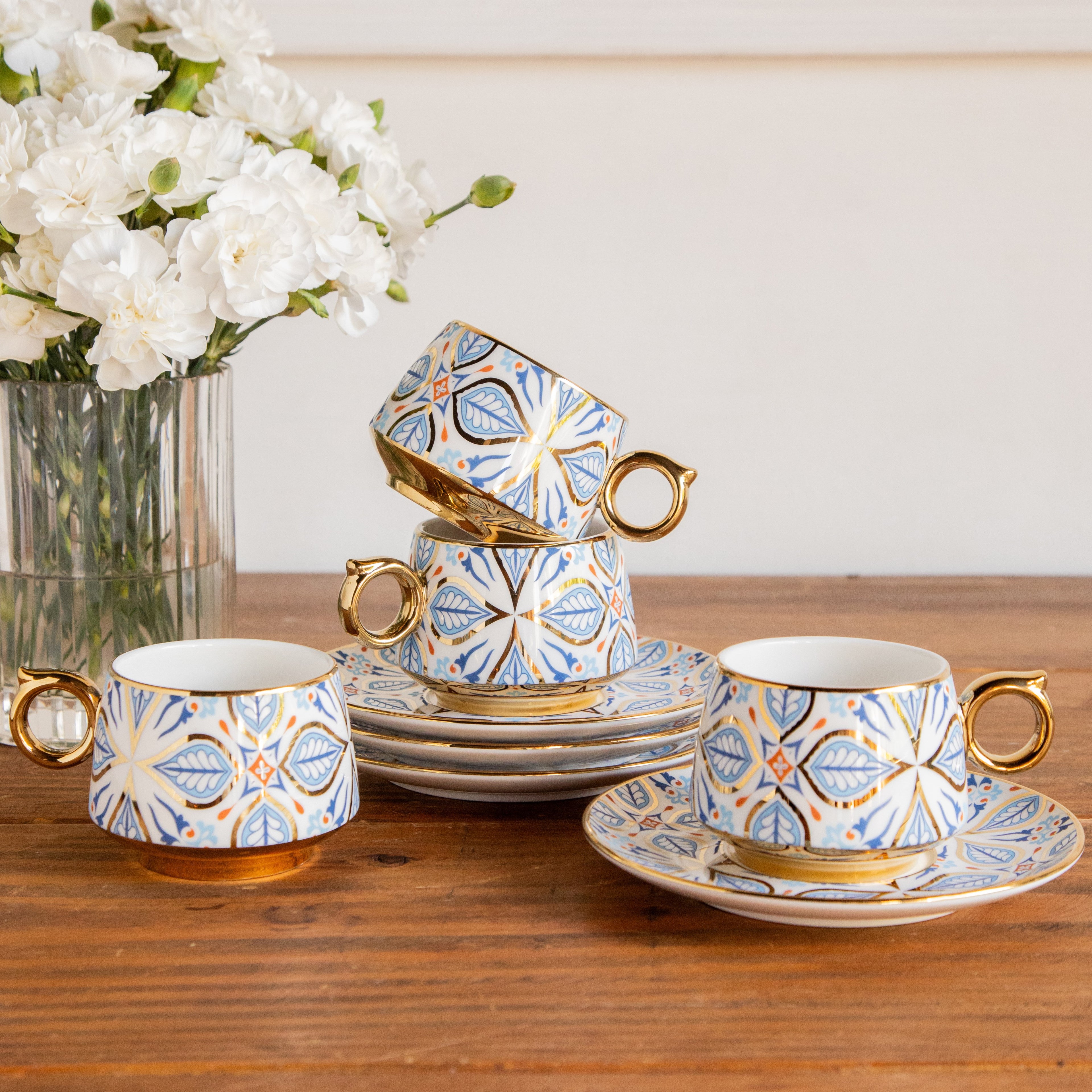 Decorative tea set with floral patterns on a wooden table, accompanied by a vase of white flowers.